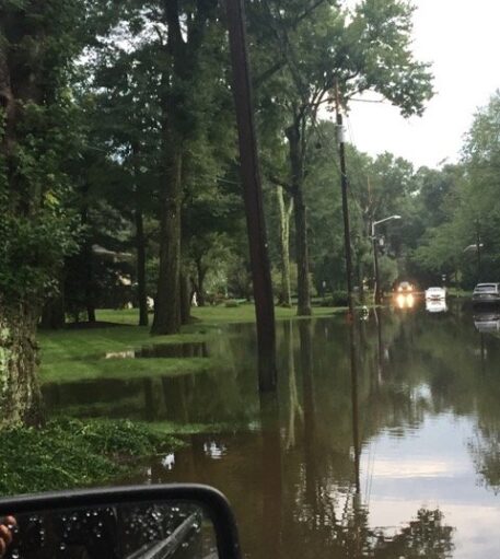 Picture of a flooded area after a storm, large trees in the background and a car avoiding the flooding.