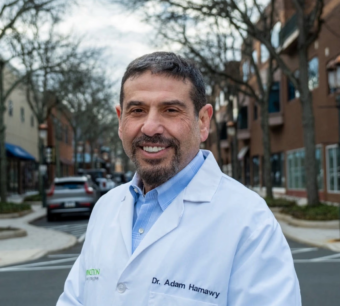 Portrait photo of Adam Hamawy in a medical jacket, with a neighborhood in the background.